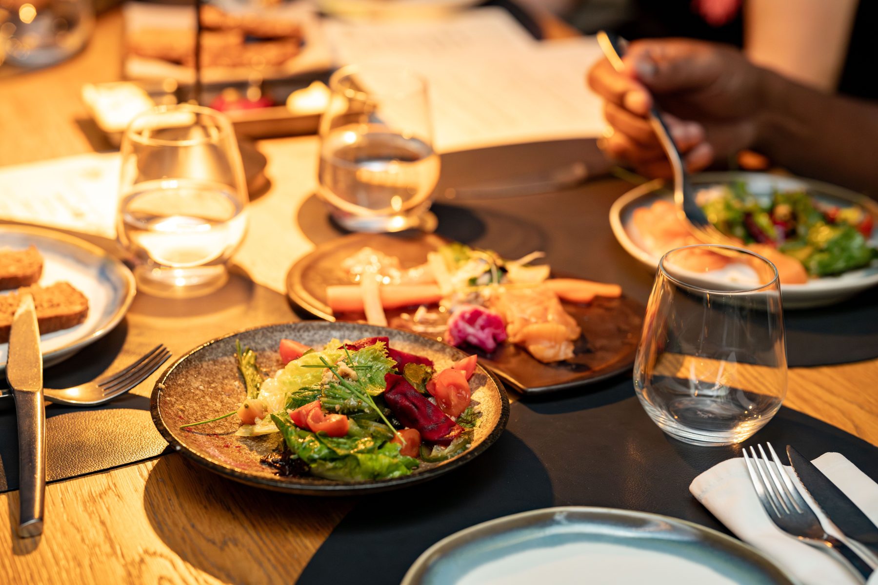 Hands sharing plates of salad and glasses of wine at a timber table