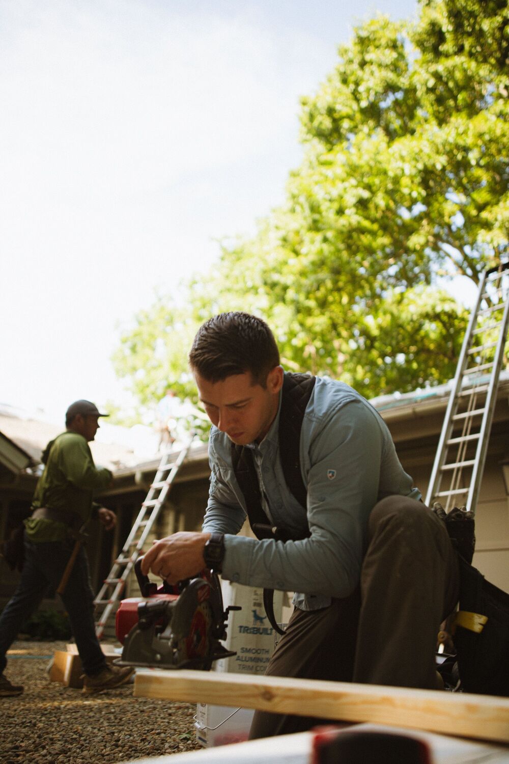 A climber crouched beside a ladder and equipment under a tall tree